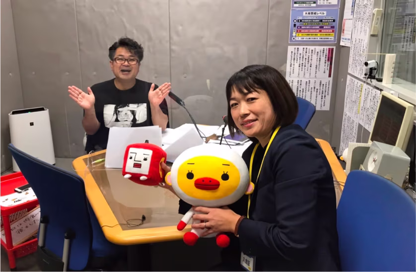 ラジオ収録中の副社長とパーソナリティがマスコットキャラクターと共にテーブルを囲んで笑顔で写っている画像