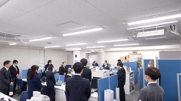 オフィス内でスーツ姿の社員たちが集まり説明を受けている様子の画像